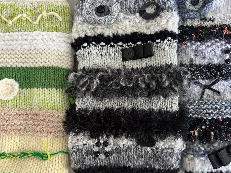 Close‑up of handmade twiddle muffs featuring textured stitches, buttons, ribbons and tactile decorations in soft, calming colours.